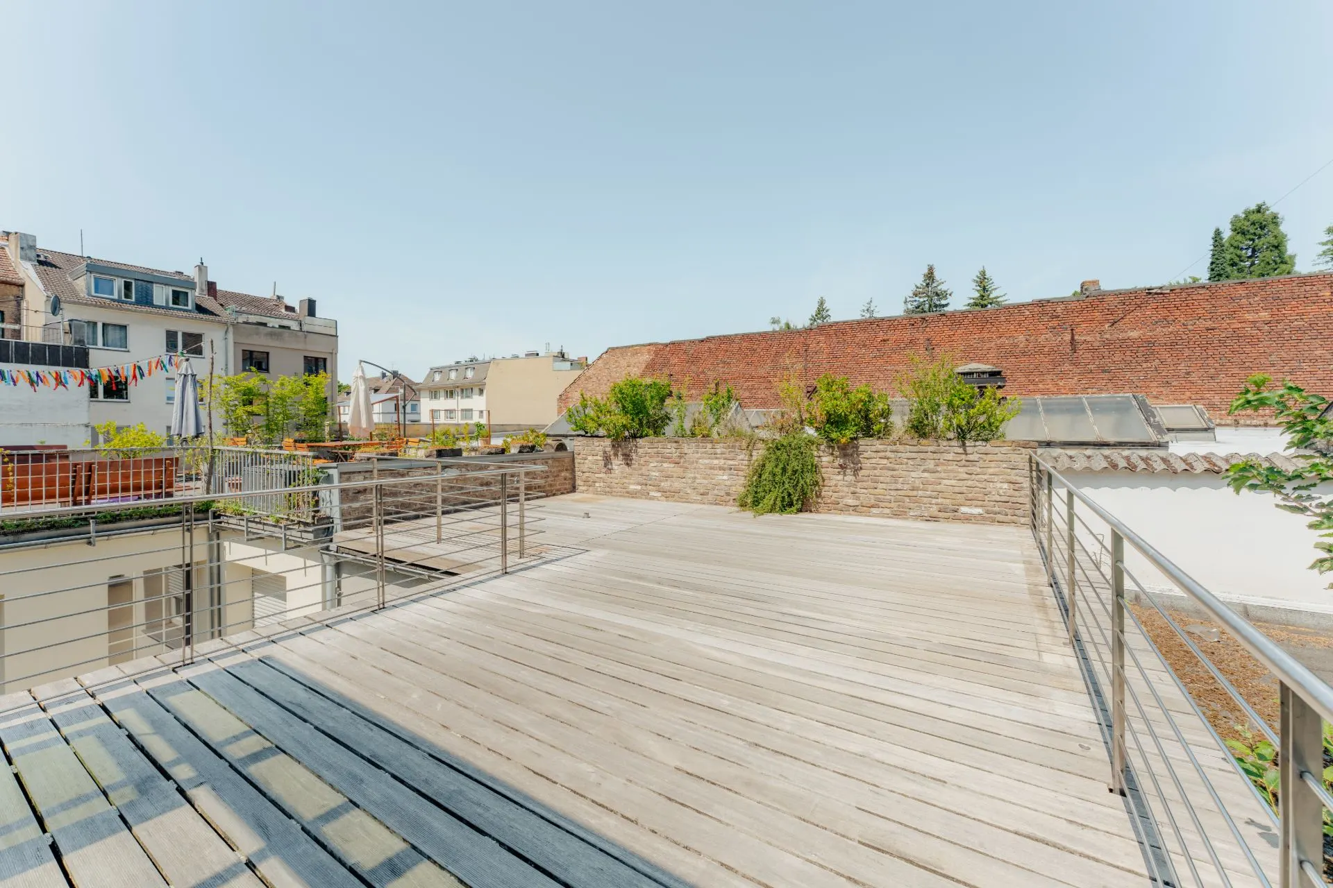 Große Holzterrasse mit Metallgeländer und Pflanzen, umgeben von Gebäuden.