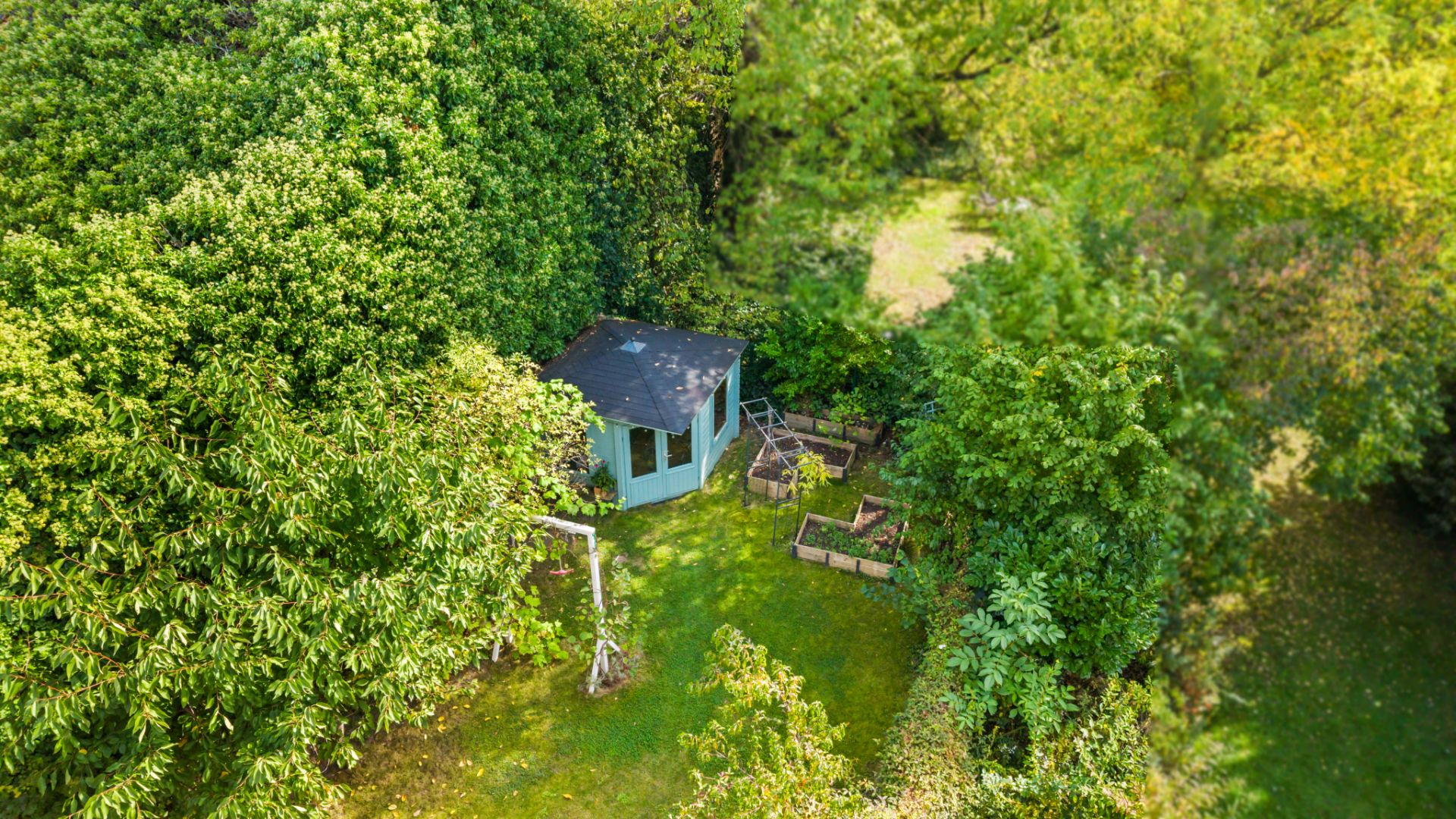 Garten mit kleinem blauen Gartenhaus, Hochbeeten und vielen Bäumen, aus der Vogelperspektive.
