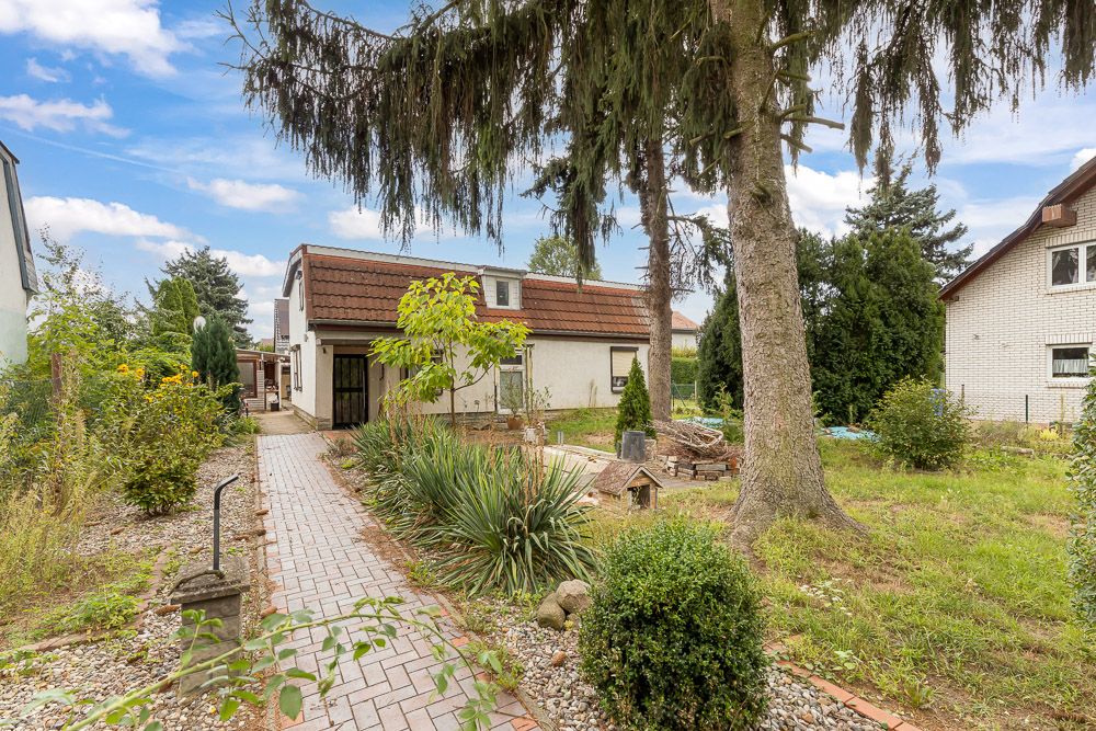 Garten mit gepflastertem Weg, Bäumen, Sträuchern und einem weißen Wohnhaus im Hintergrund.
