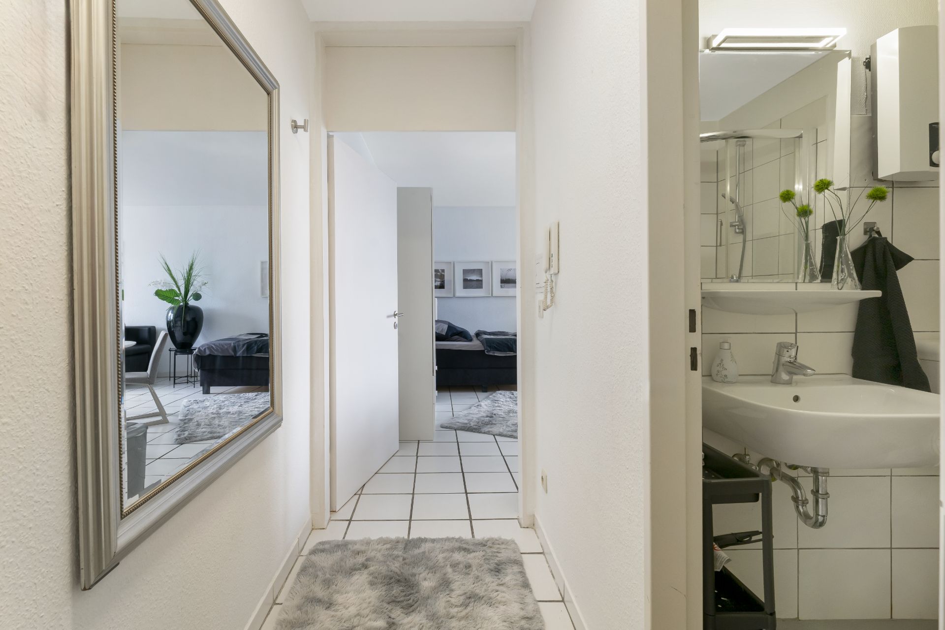 Heller Flur mit Teppich, großem Spiegel, Blick ins Wohnzimmer und Badezimmer mit Waschbecken.