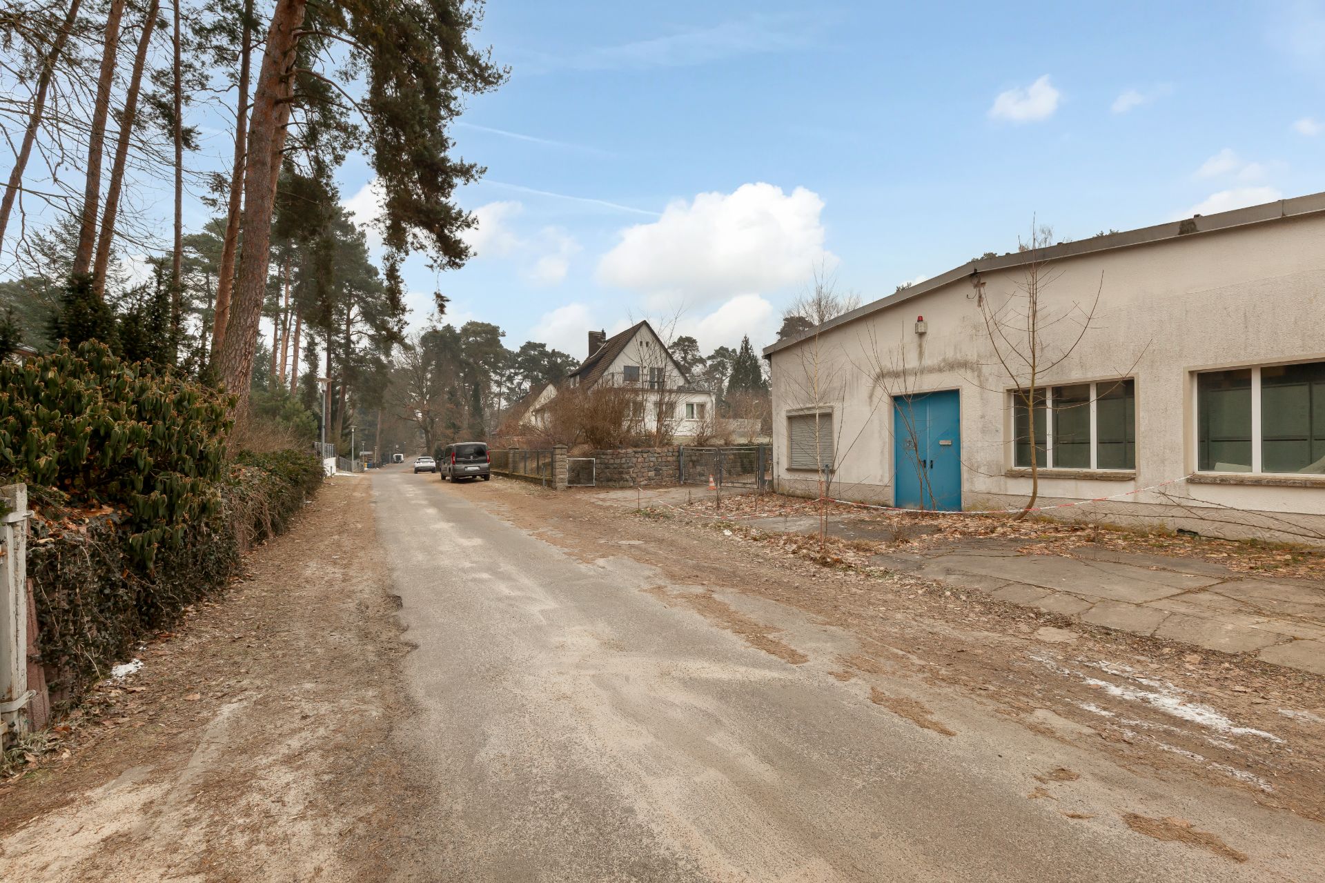Schmale Straße mit Bäumen, einem Wohnhaus und einem flachen Gebäude mit blauer Tür.