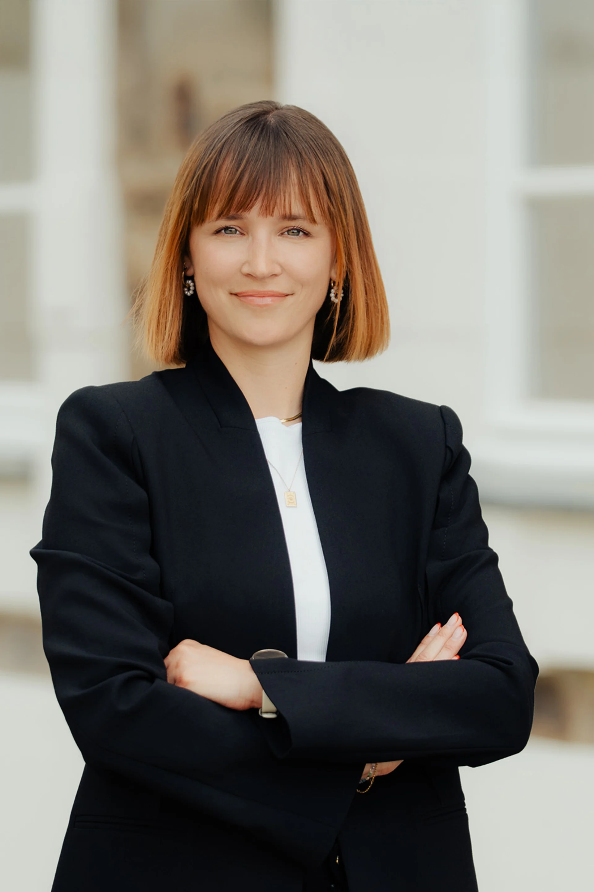 Eine Frau mit schulterlangem Haar trägt einen dunklen Blazer und steht mit verschränkten Armen vor einem Fenster.