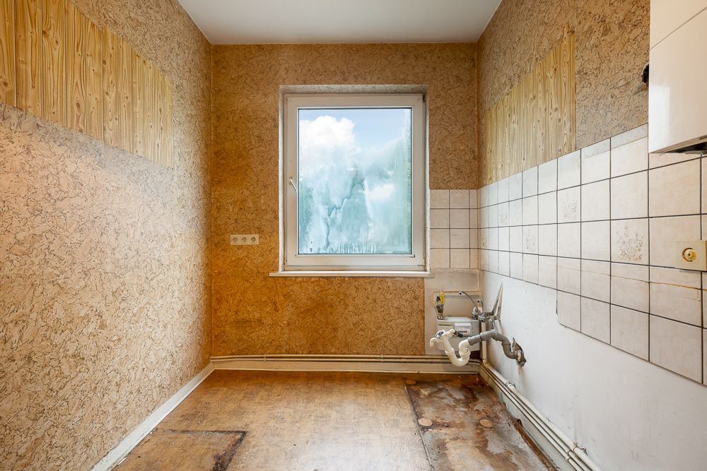 Leerer Raum mit brauner Tapete, einem Fenster und sichtbaren Rohrleitungen an der Wand.
