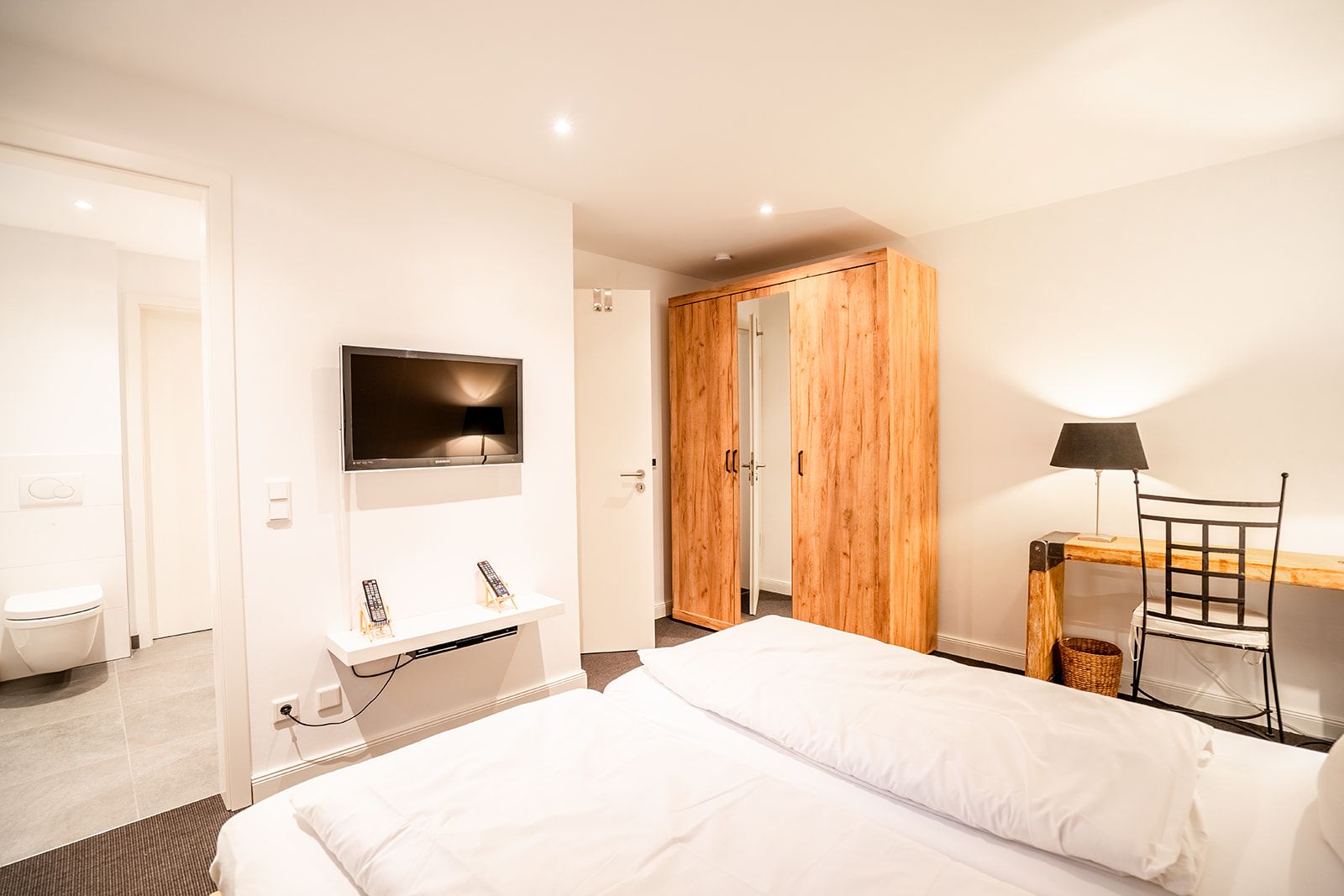 Helles Schlafzimmer mit Doppelbett, Holzschrank, Schreibtisch, Stuhl, Fernseher und Zugang zum Badezimmer.
