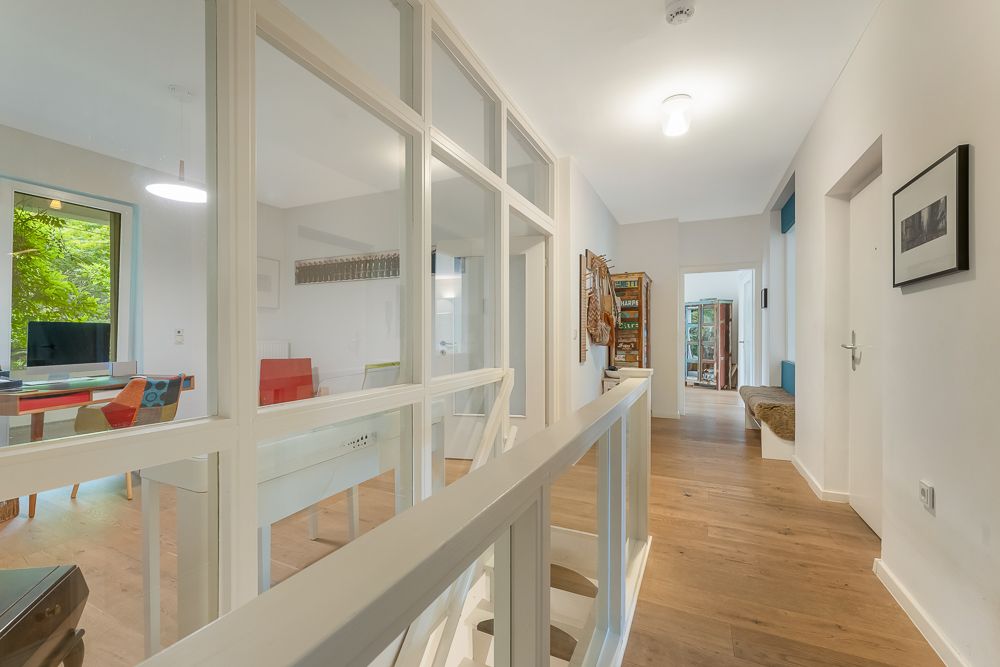 Heller Flur mit Holzboden, weißen Wänden und großen Glaswänden zu einem Arbeitszimmer.