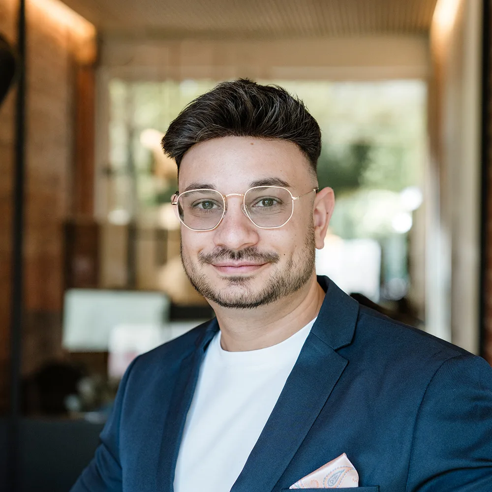 Ein Mann mit Brille und Anzug steht in einem modernen, hellen Raum.