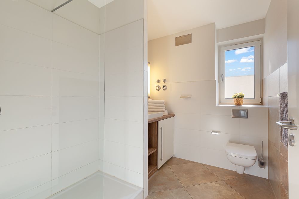 Modernes Badezimmer mit bodengleicher Dusche, WC, Waschbecken und Fenster.