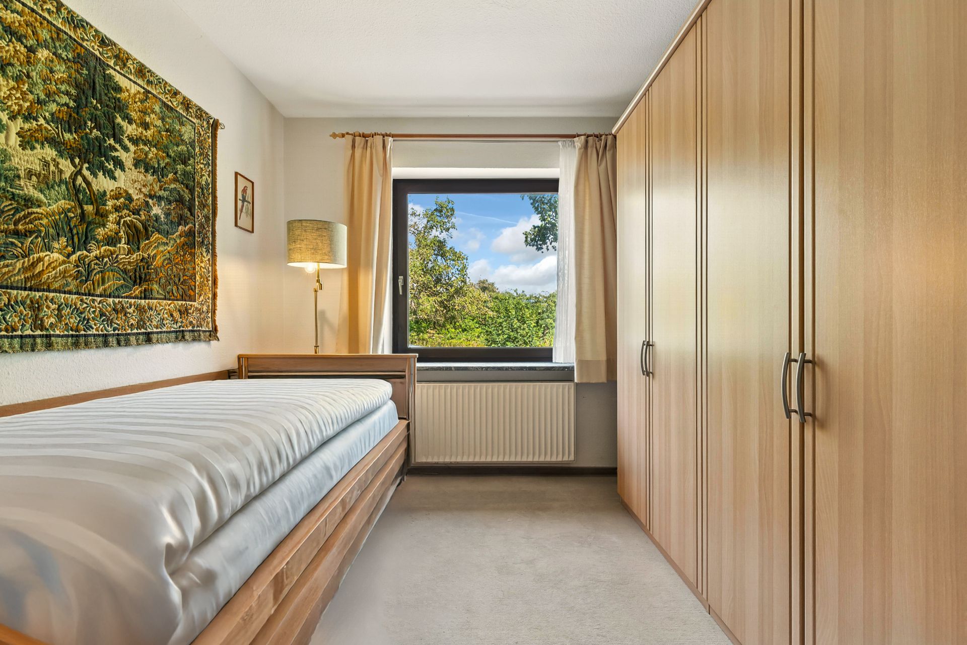 Schlafzimmer mit Einzelbett, großem Holzschrank und Fenster mit Blick ins Grüne.