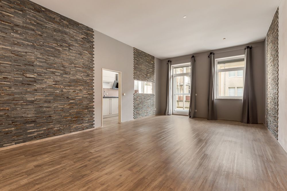 Leeres Zimmer mit Holzfußboden, Steinwänden, großen Fenstern und Vorhängen.