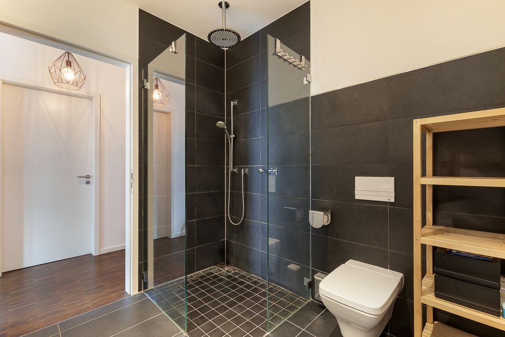 Modernes Badezimmer mit begehbarer Dusche, WC und Holzregal, dunkle Fliesen.