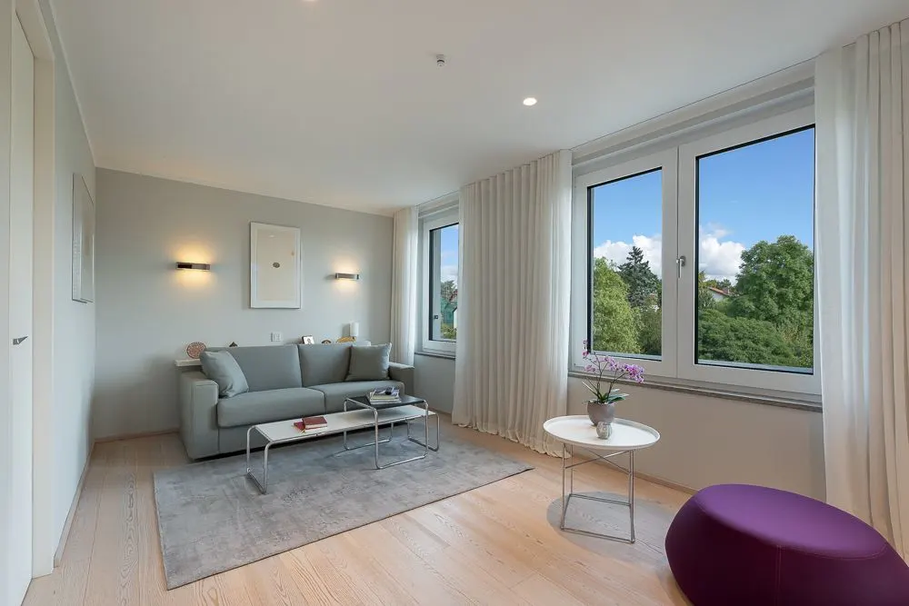 Helles Wohnzimmer mit grauem Sofa, Couchtisch, runder Beistelltisch und großen Fenstern mit Blick ins Grüne.