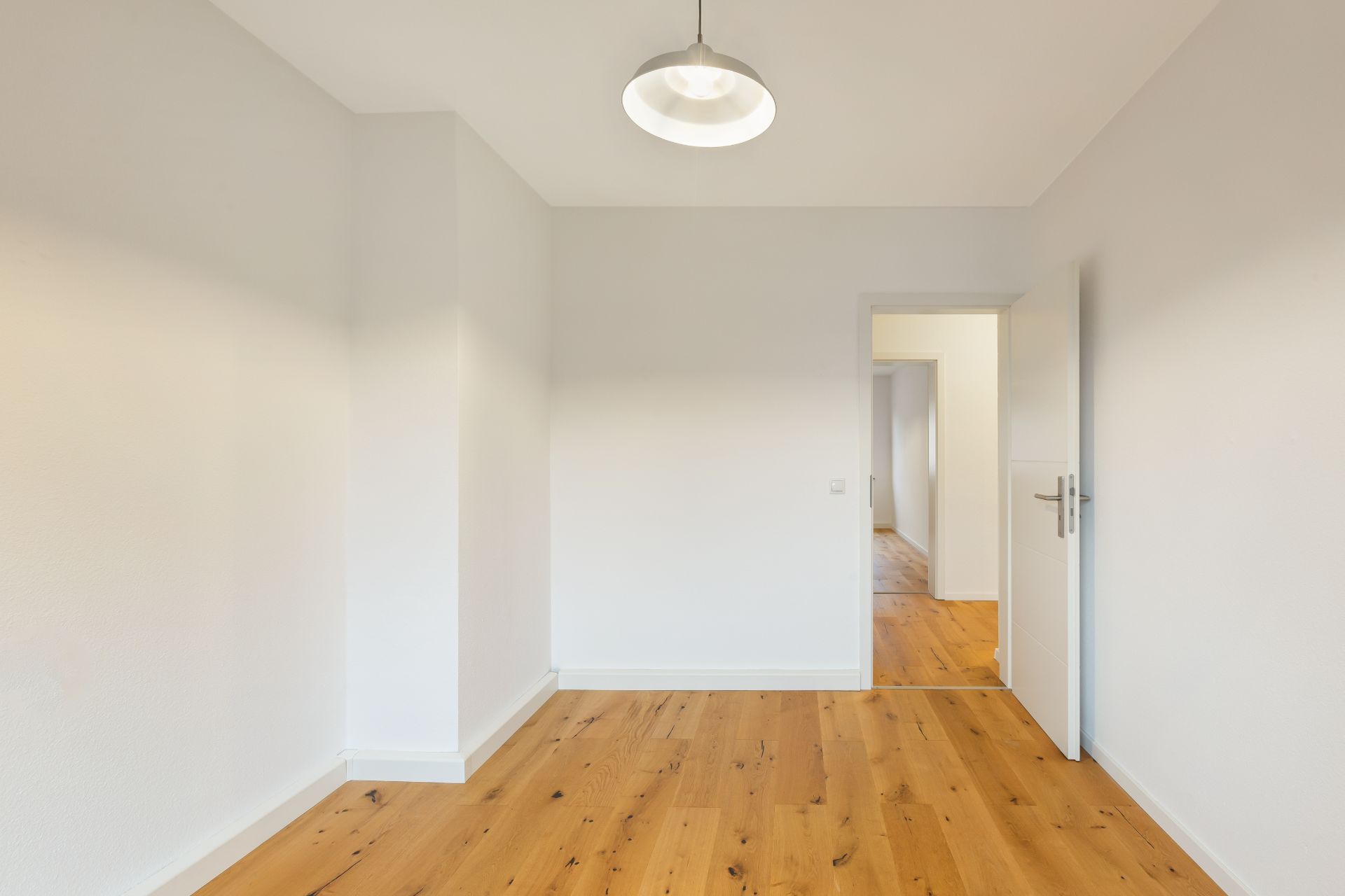 Leeres Zimmer mit Holzboden, weißen Wänden, offener Tür und Deckenlampe.