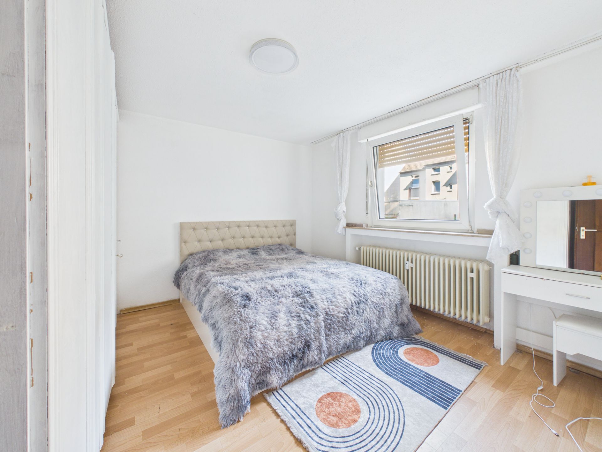 Helles Schlafzimmer mit Bett, Fenster, Teppich und Schminktisch auf Holzfußboden.