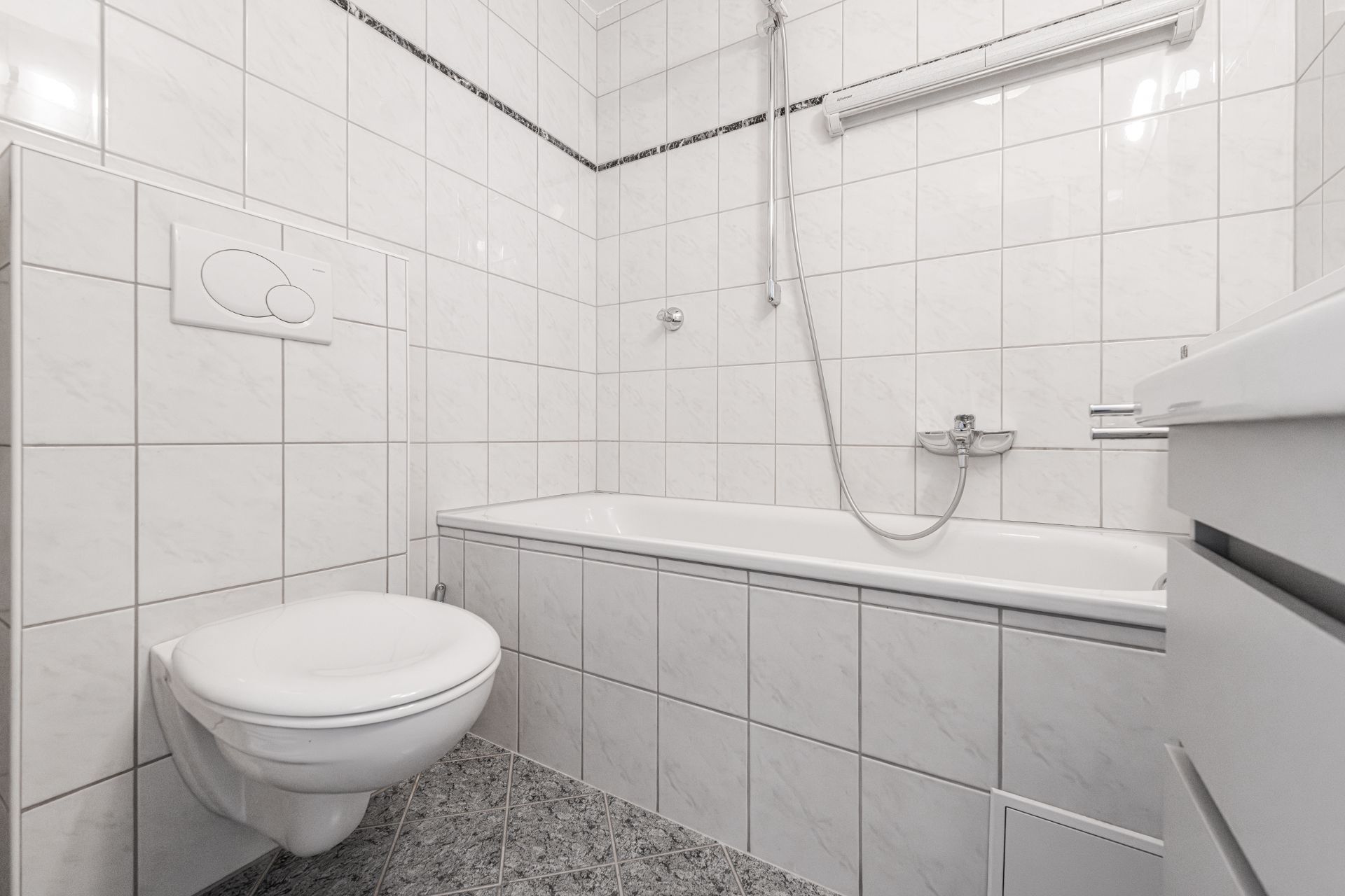 Helles Bad mit Wandtoilette, Badewanne, Handbrause und weißen Fliesen.