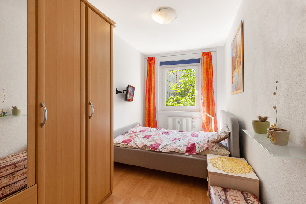Kleines Schlafzimmer mit Bett, Kleiderschrank, Fenster und orangefarbenen Vorhängen.