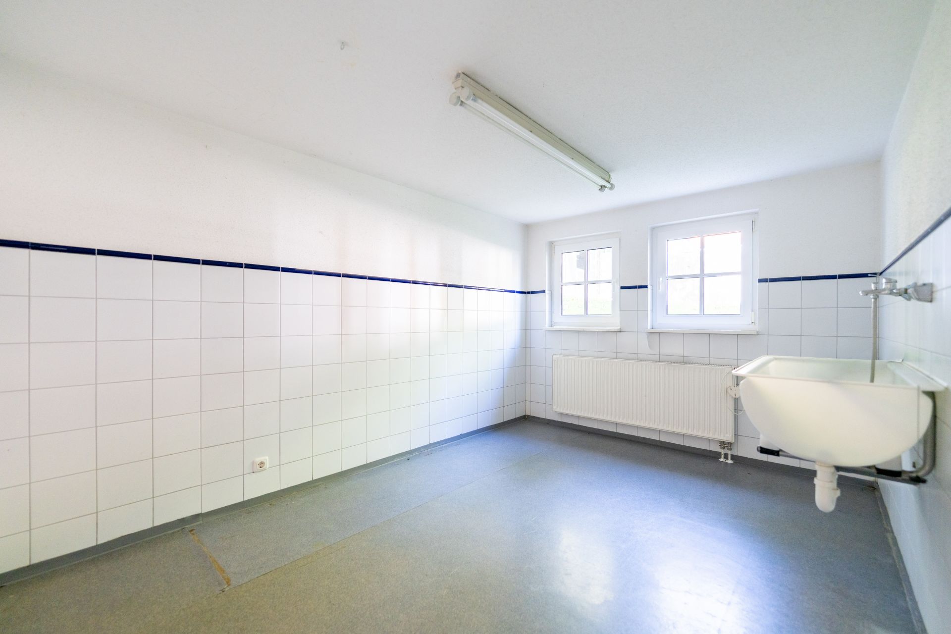 Heller, leerer Raum mit weißen Wandfliesen, zwei Fenstern und einem Waschbecken an der Wand.