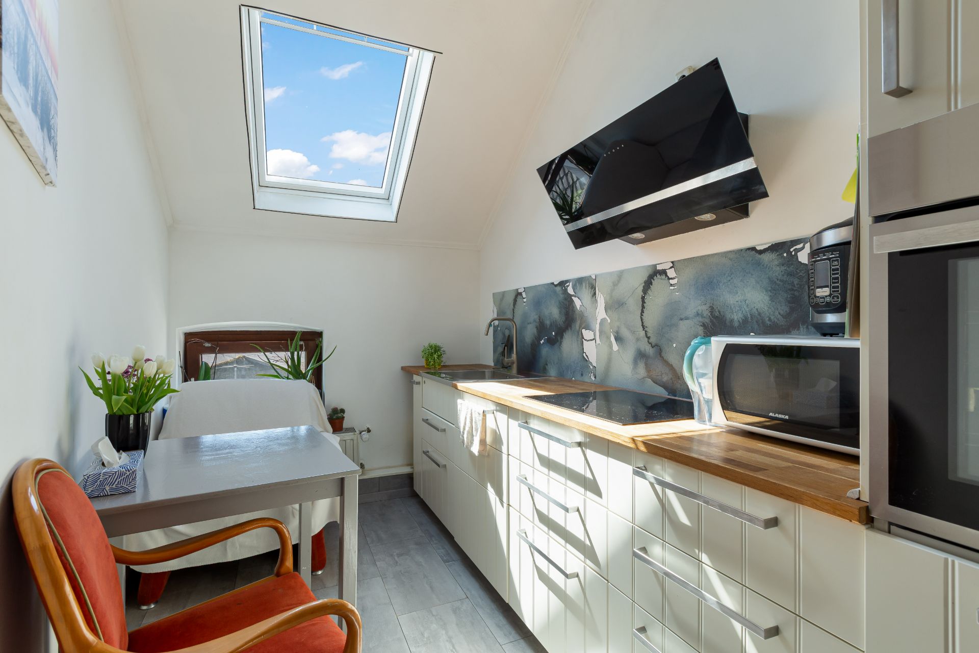 Helle Küche mit Dachfenster, Holztisch, Stuhl, Pflanzen und moderner Küchenausstattung.