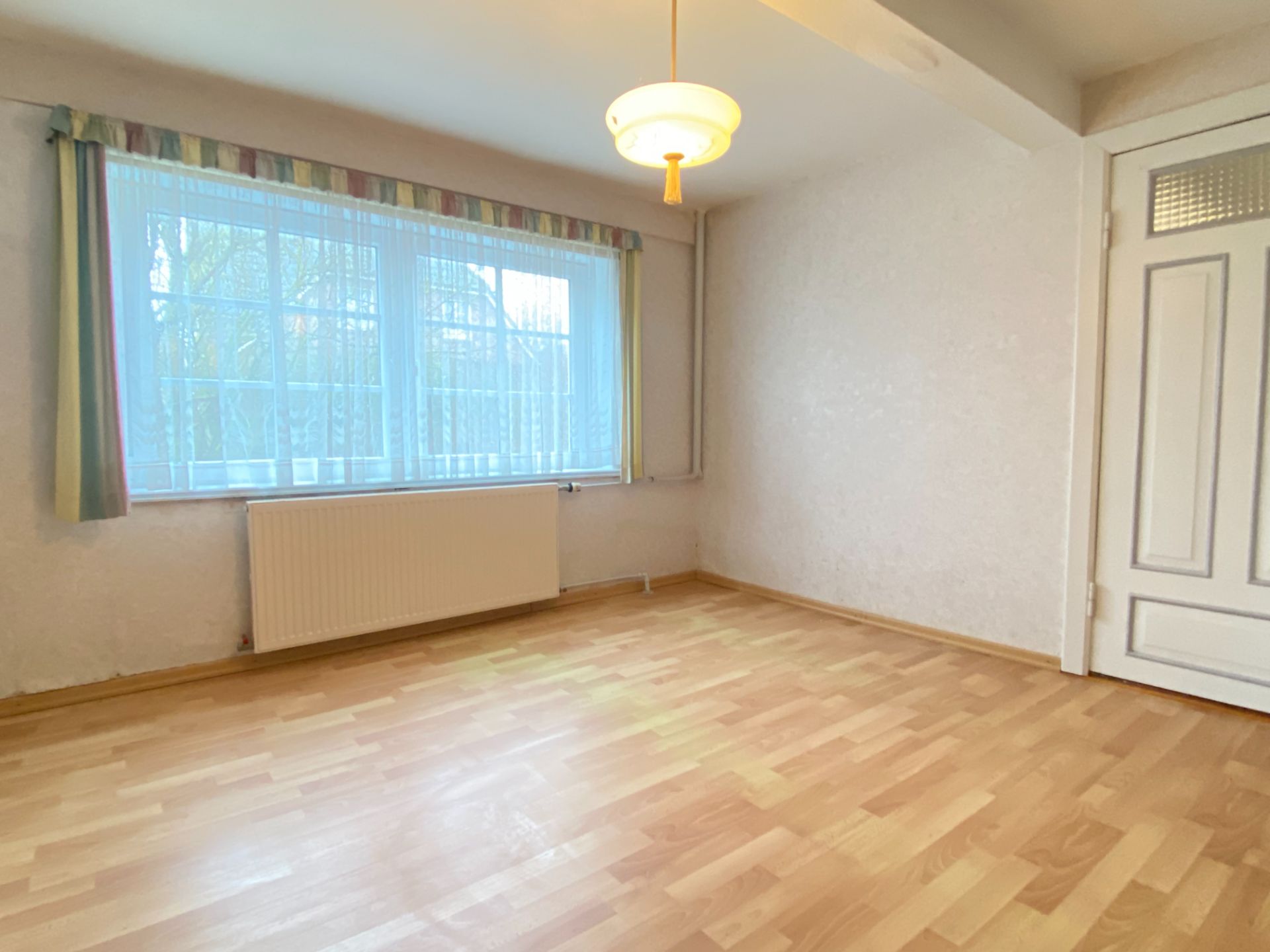 Leerer Raum mit Holzfußboden, großem Fenster mit Vorhang, Heizung und weißer Tür.