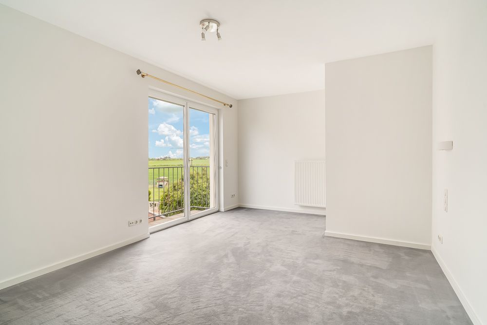 Helles, leeres Zimmer mit Teppichboden und großer Glastür zum Balkon, Blick ins Grüne.