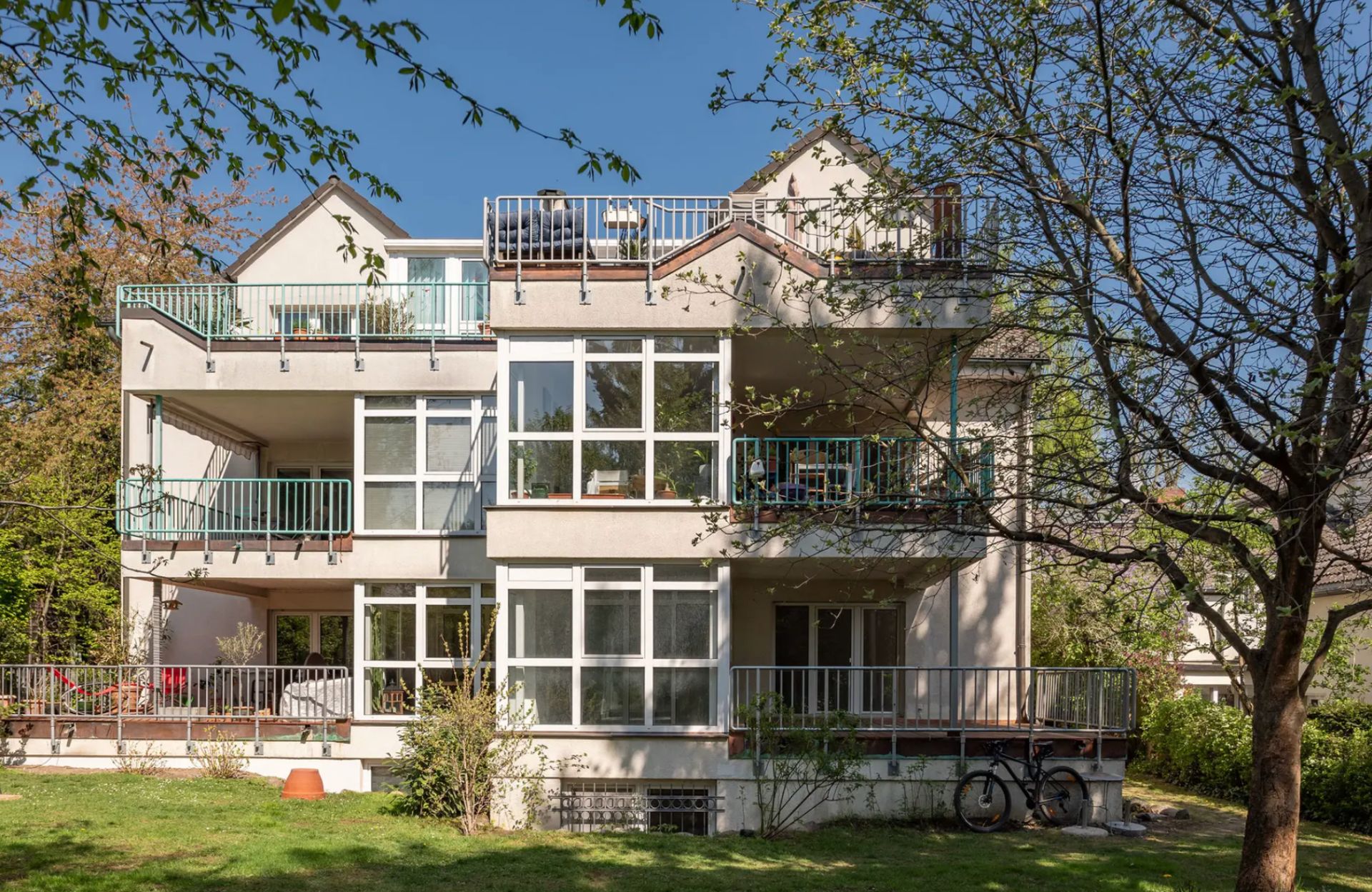 Modernes Wohnhaus mit mehreren Balkonen und großen Fenstern, umgeben von Bäumen und Garten.
