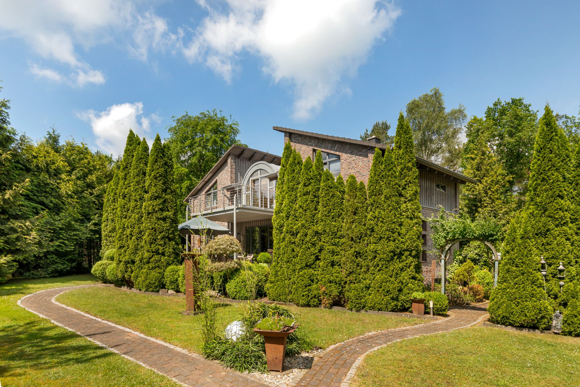 Backsteinhaus mit Balkon, umgeben von hohen Bäumen und gepflegtem Garten.