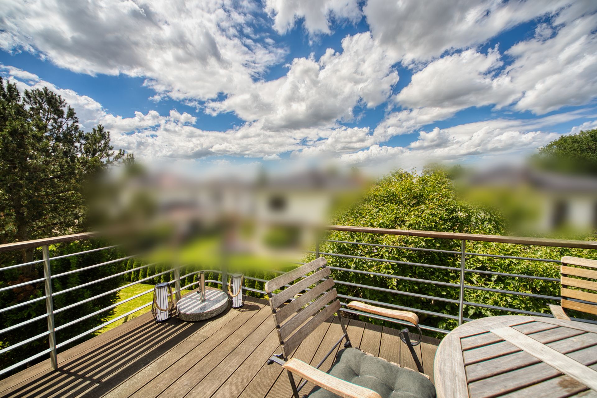 Balkon mit Holzboden, Tisch, Stühlen und Ausblick auf Bäume unter blauem Himmel mit Wolken.