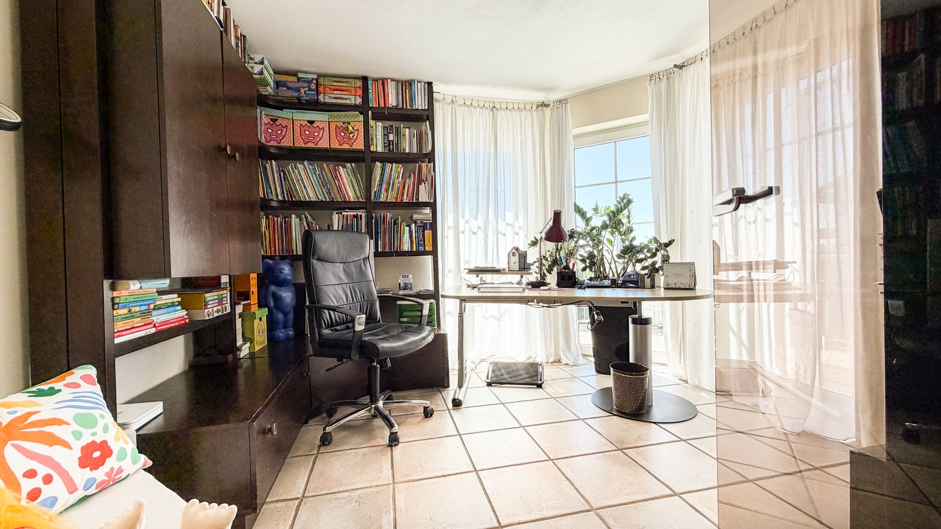 Helles Arbeitszimmer mit Bücherregal, Schreibtisch, Bürostuhl und großen Fenstern mit Vorhängen.
