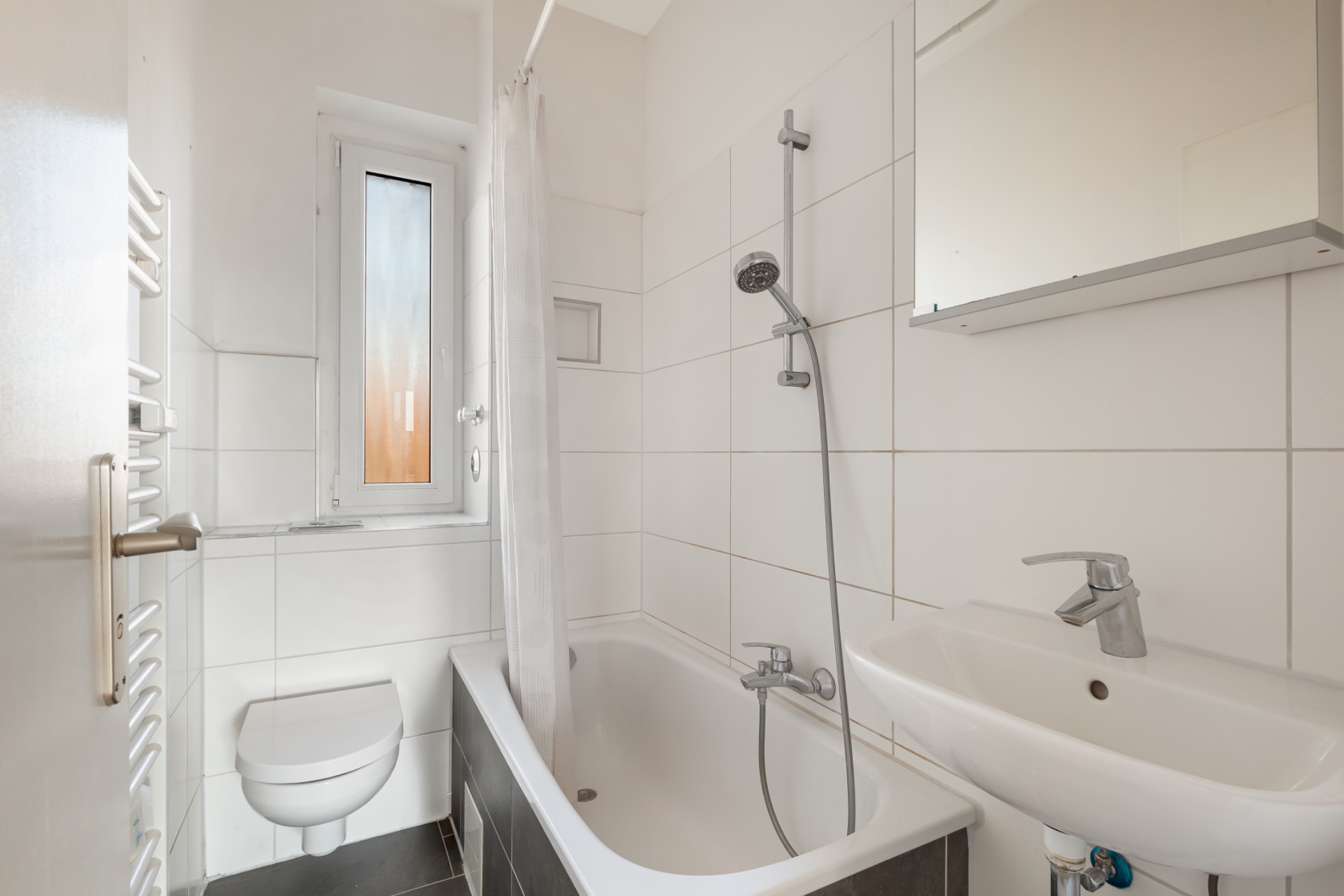 Kleines Badezimmer mit Badewanne, Duschvorhang, Wandtoilette, Waschbecken und Fenster.