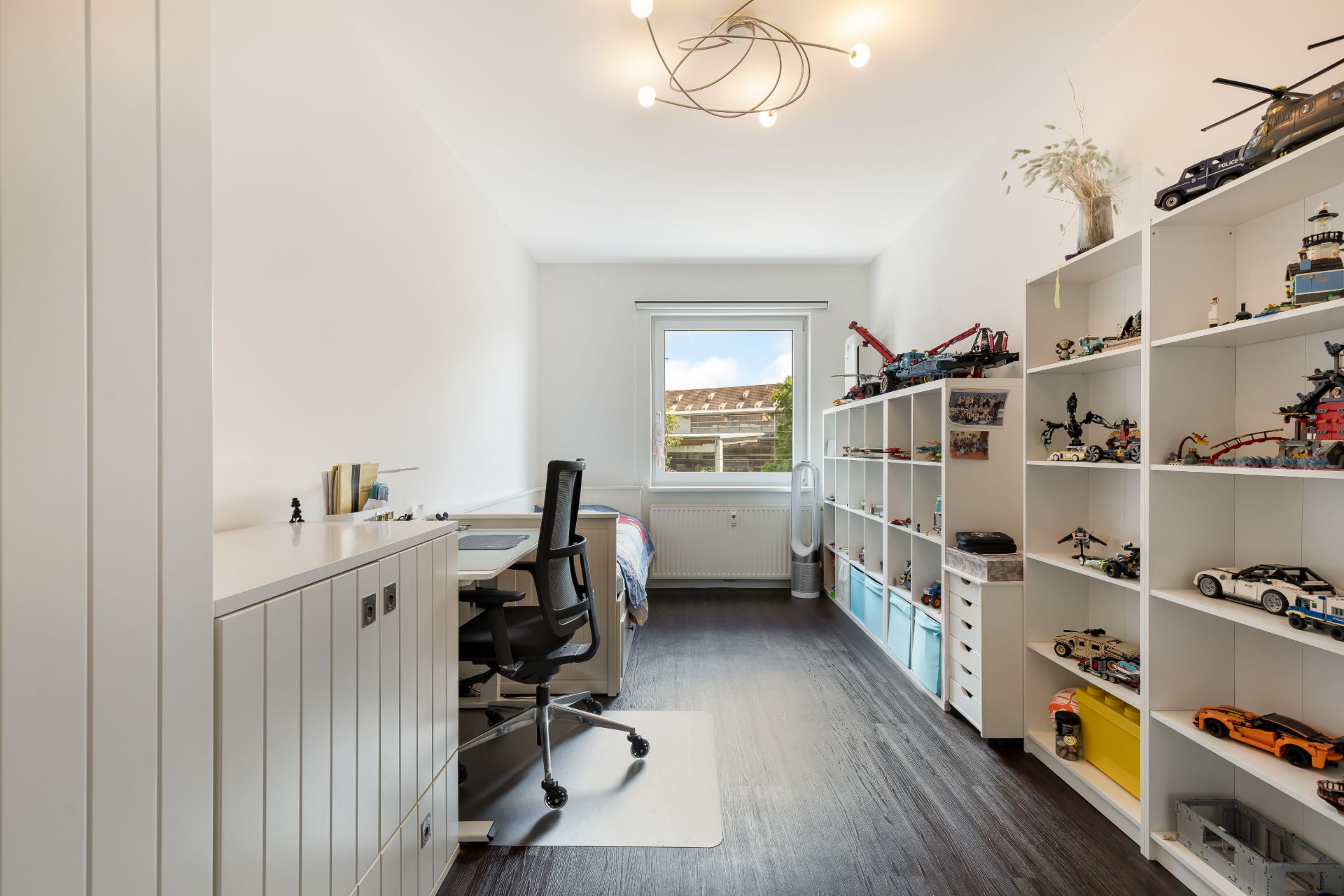 Helles Kinderzimmer mit Schreibtisch, Regal voller Spielzeug und einem Fenster im Hintergrund.