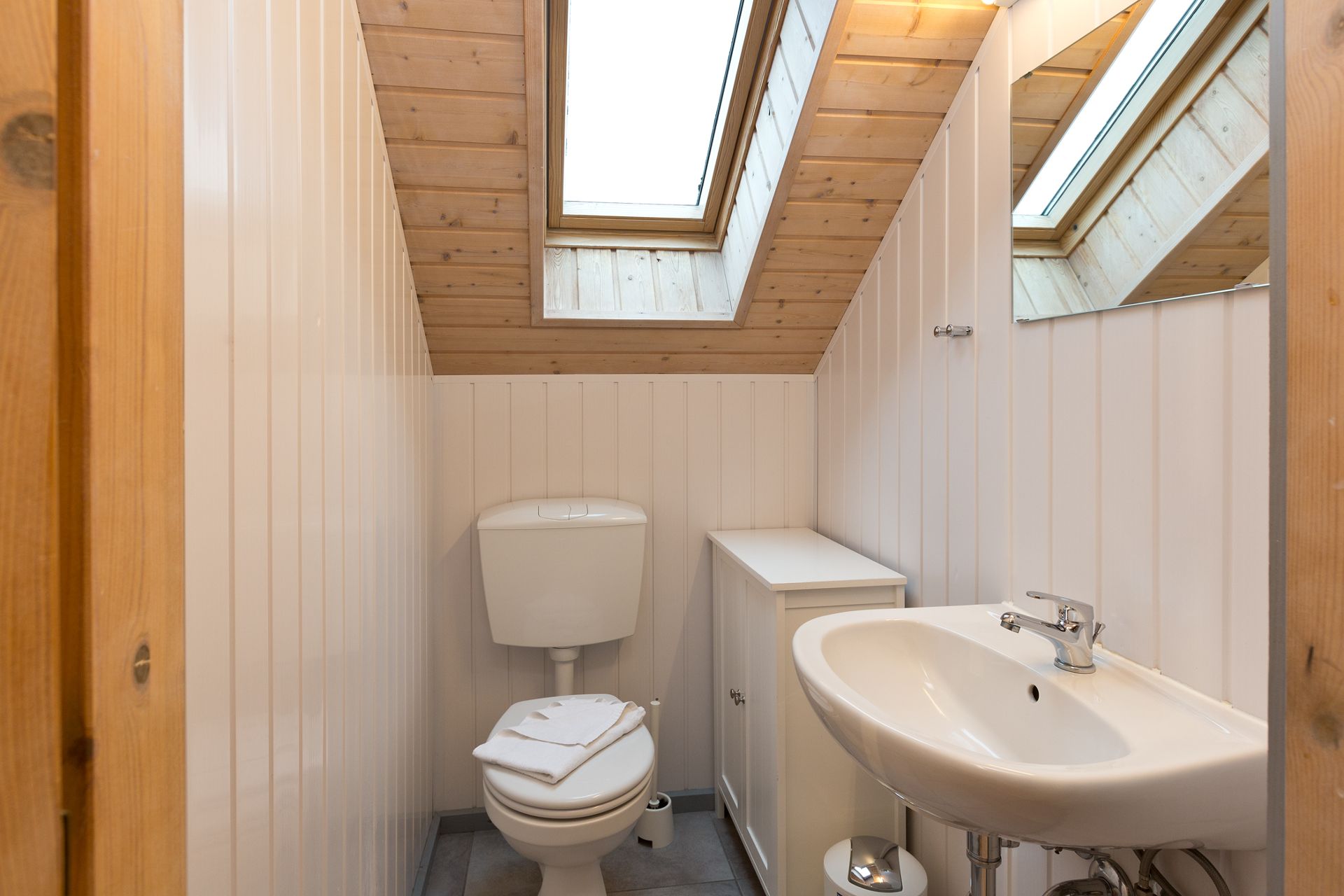 Kleines Bad mit Toilette, Waschbecken und Dachfenster, weiße Vertäfelung und Holzdecke.