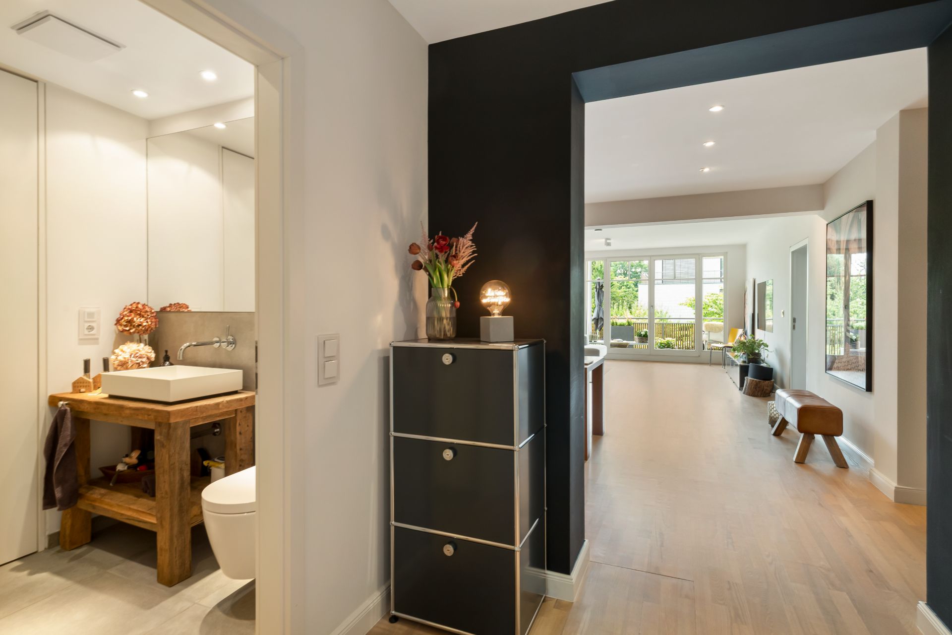 Flur mit Blick ins Wohnzimmer, links ein Bad mit Holzwaschtisch und Spiegel, hell und modern eingerichtet.