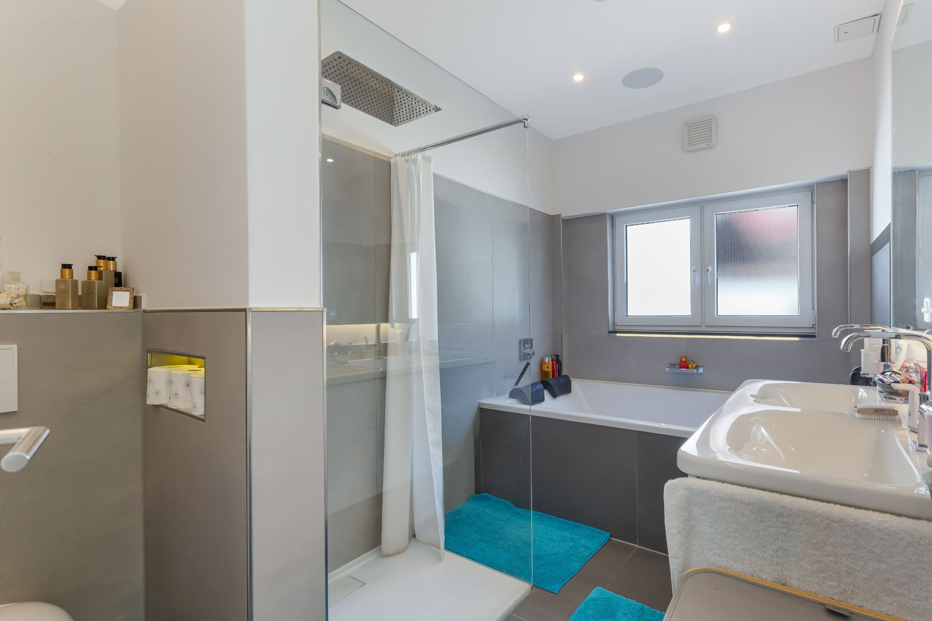 Modernes Badezimmer mit Dusche, Badewanne, Doppelwaschbecken und zwei blauen Teppichen.