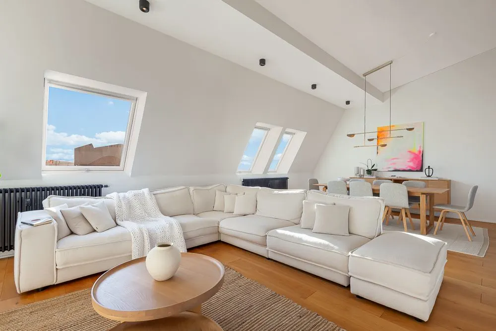 Helles Wohnzimmer mit weißem Sofa, rundem Holztisch und Essbereich im Hintergrund.