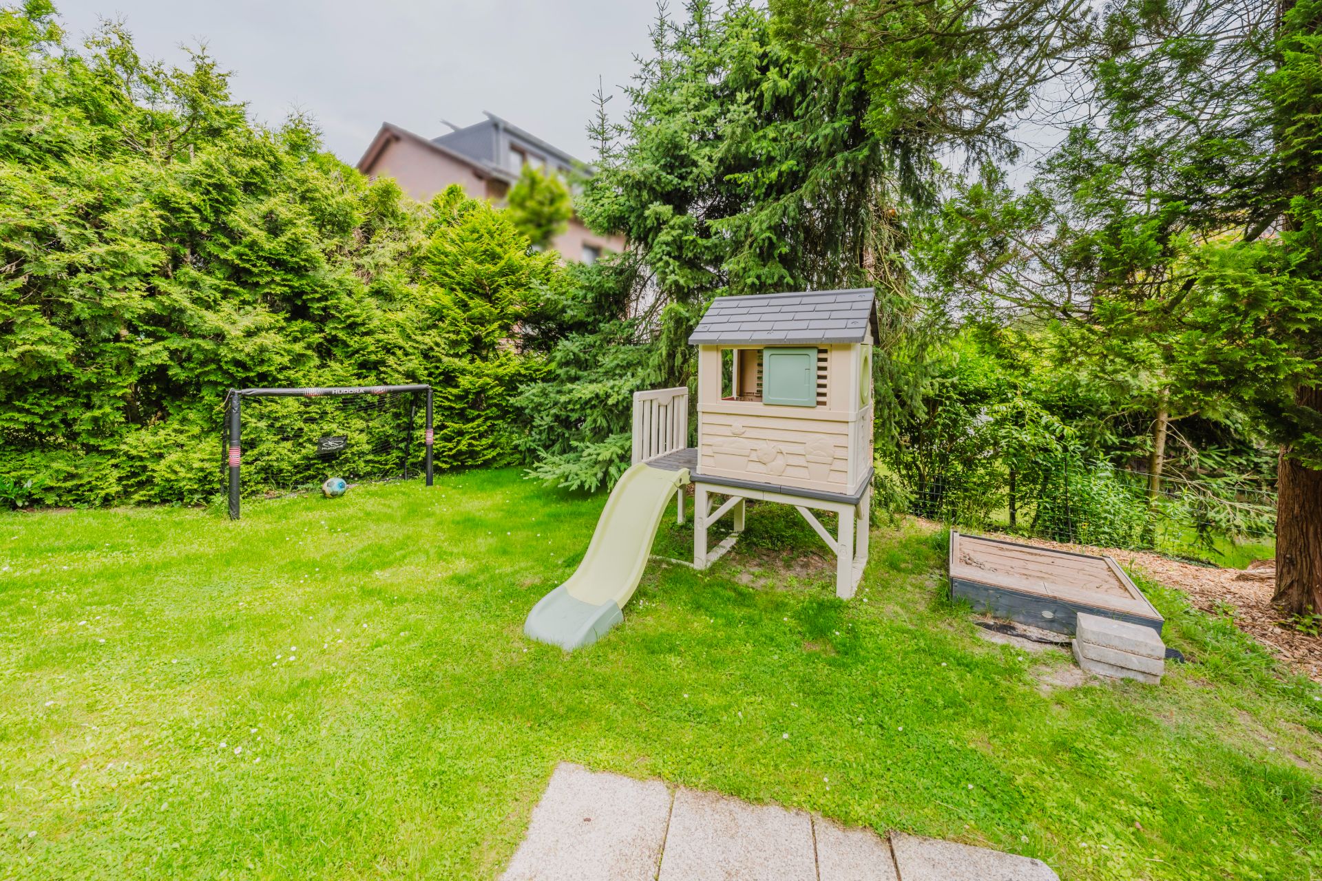 Garten mit Spielhaus, Rutsche, Sandkasten und kleinem Fußballtor auf grünem Rasen.
