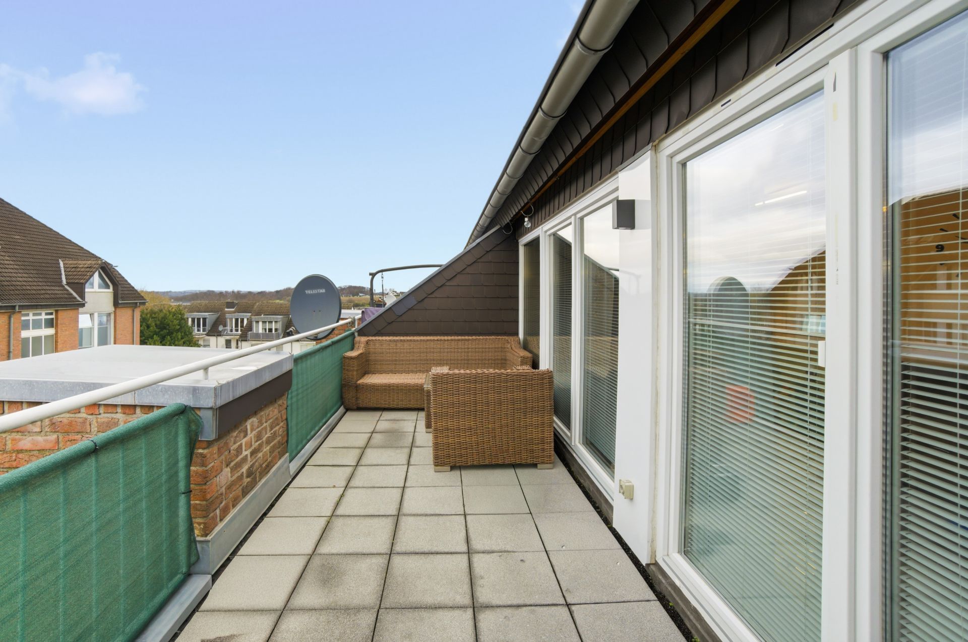 Großer Balkon mit gefliestem Boden, Rattan-Sitzgruppe und Blick auf Nachbarhäuser.
