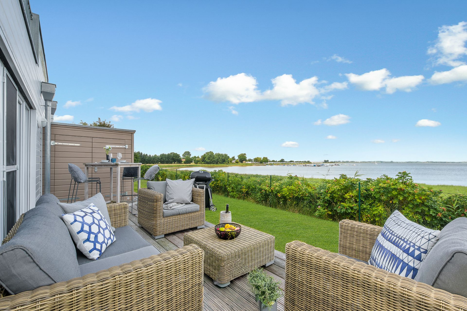 Terrasse mit Sitzmöbeln, Tisch und Blick auf einen See und Wiese bei klarem Himmel.