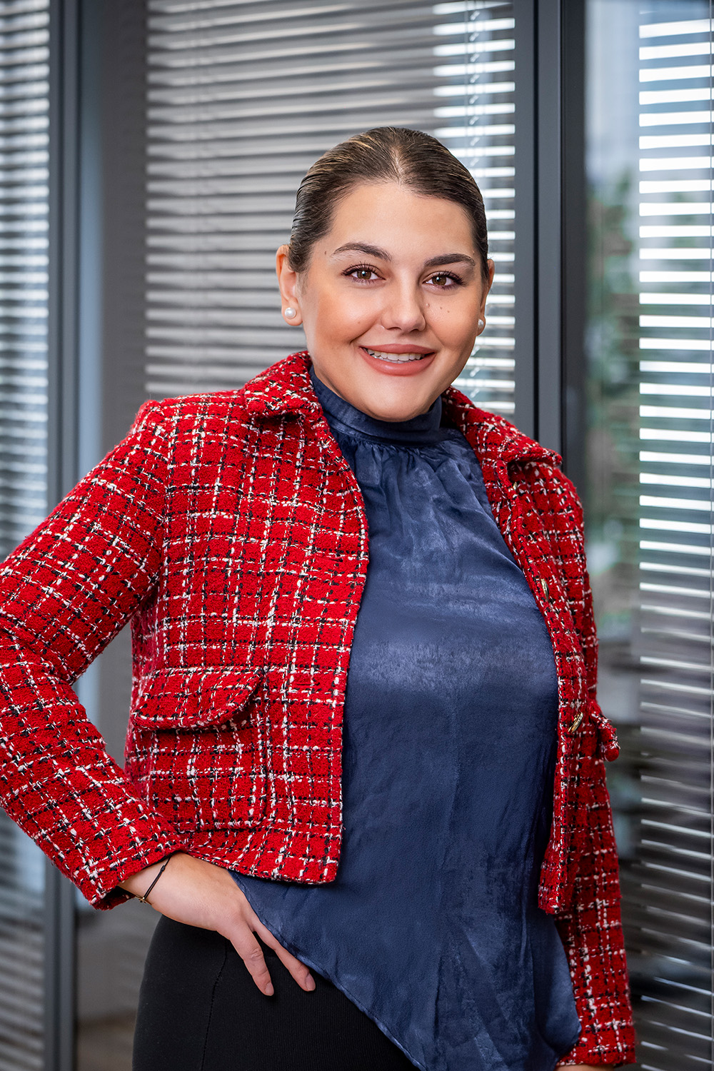 Frau mit rotem Blazer steht vor Fenstern mit Jalousien.