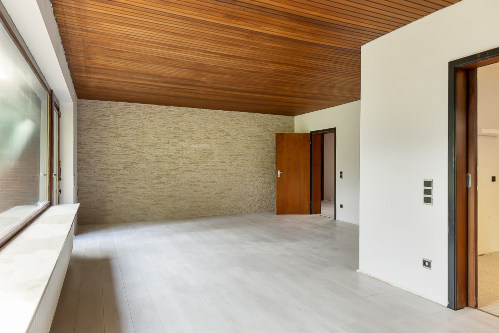 Leerer Raum mit Holzdecke, heller Wand, großem Fenster und offener Tür.