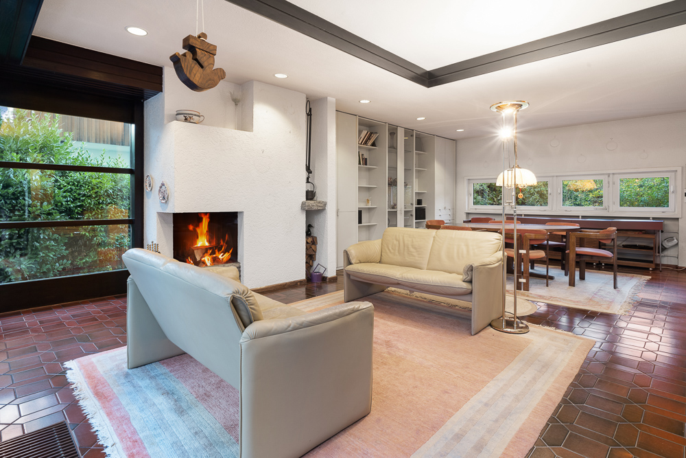 Wohnzimmer mit zwei Sofas, Kamin, Esstisch und großen Fenstern, die auf einen Garten blicken.