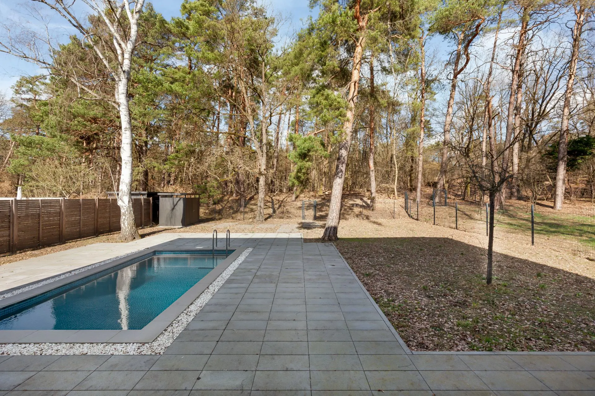 Gepflasterte Terrasse mit rechteckigem Pool, angrenzend an einen Wald mit Bäumen.