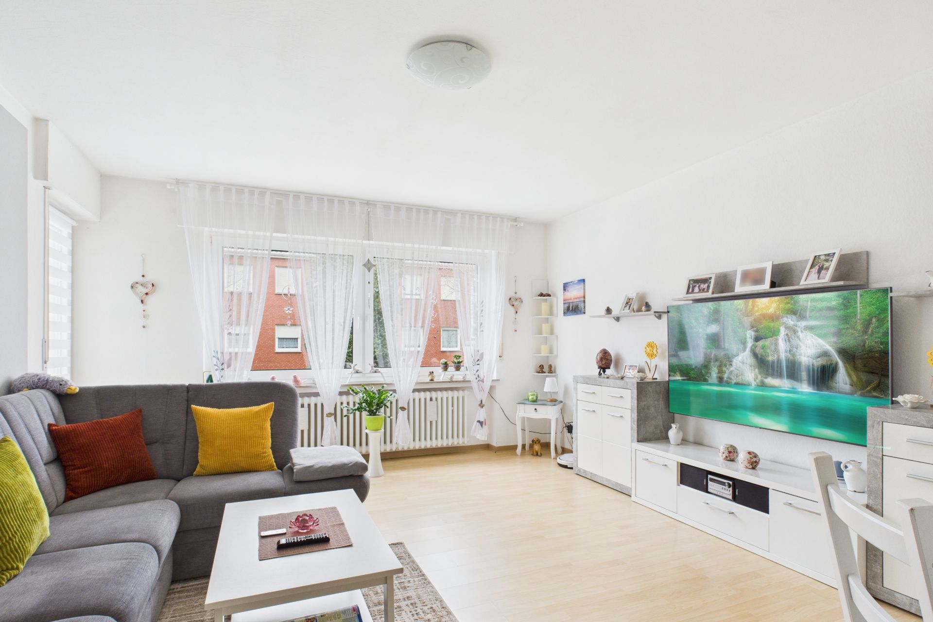 Helles Wohnzimmer mit grauem Sofa, Couchtisch, TV und großen Fenstern mit weißen Vorhängen.