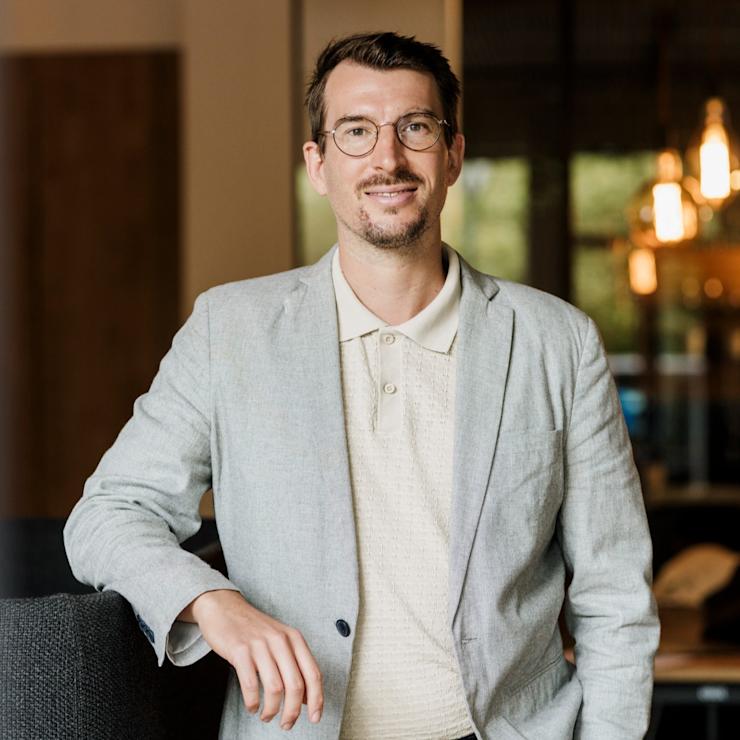 Mann mit Brille und hellem Sakko steht mit verschränkten Armen vor einer Wand.