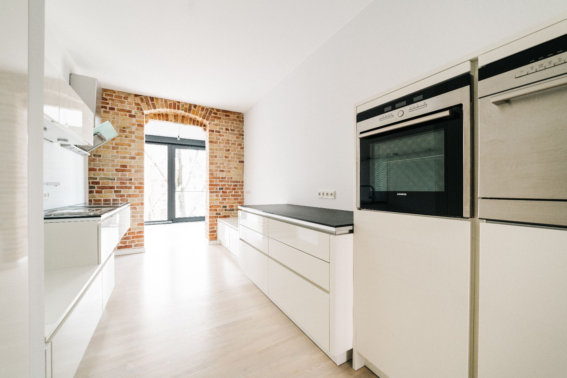 Moderne weiße Küche mit Backofen, Kochfeld und Backsteinwand am Fenster.