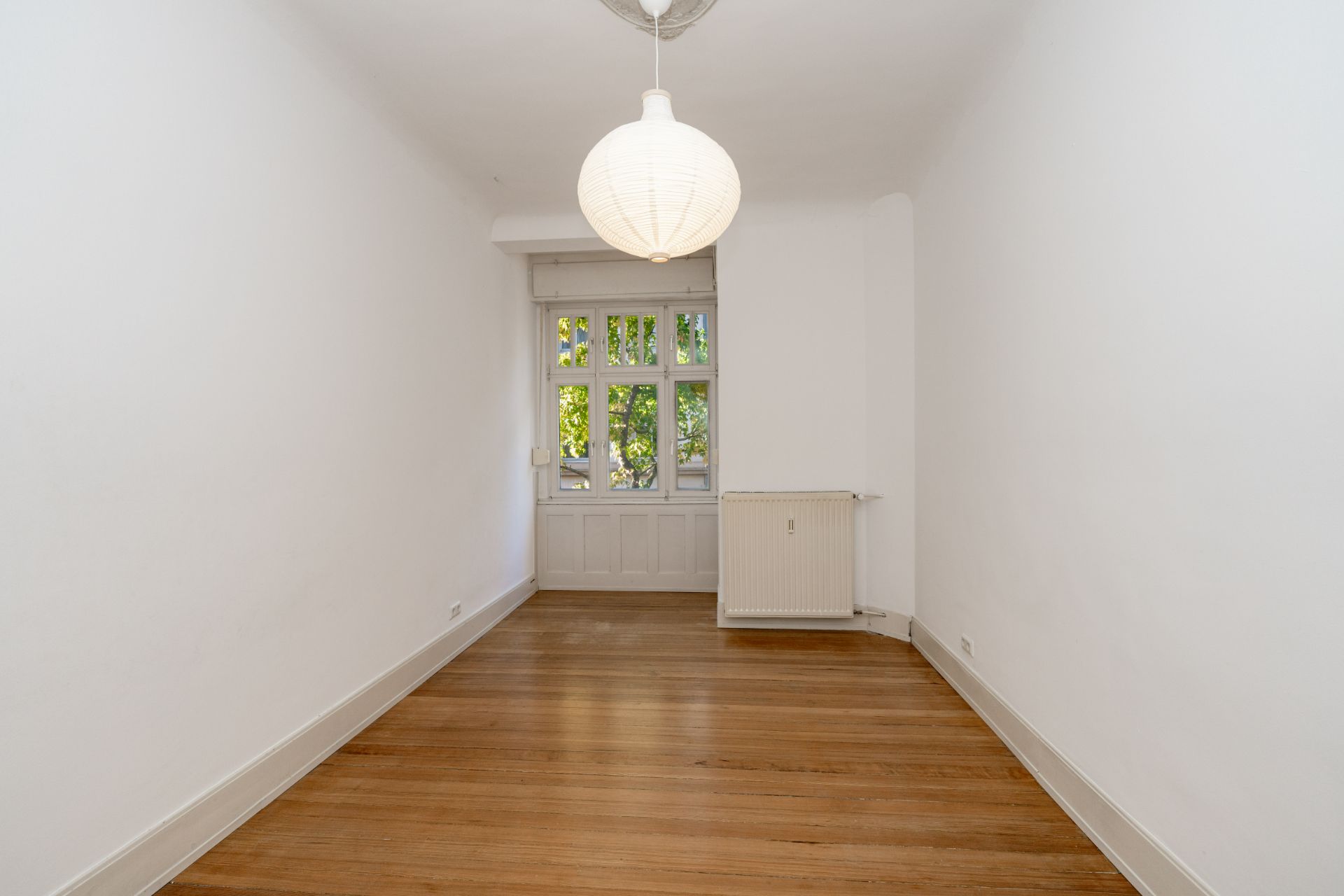 Leerer Raum mit Holzboden, weiß gestrichenen Wänden, Fenster und Hängeleuchte.