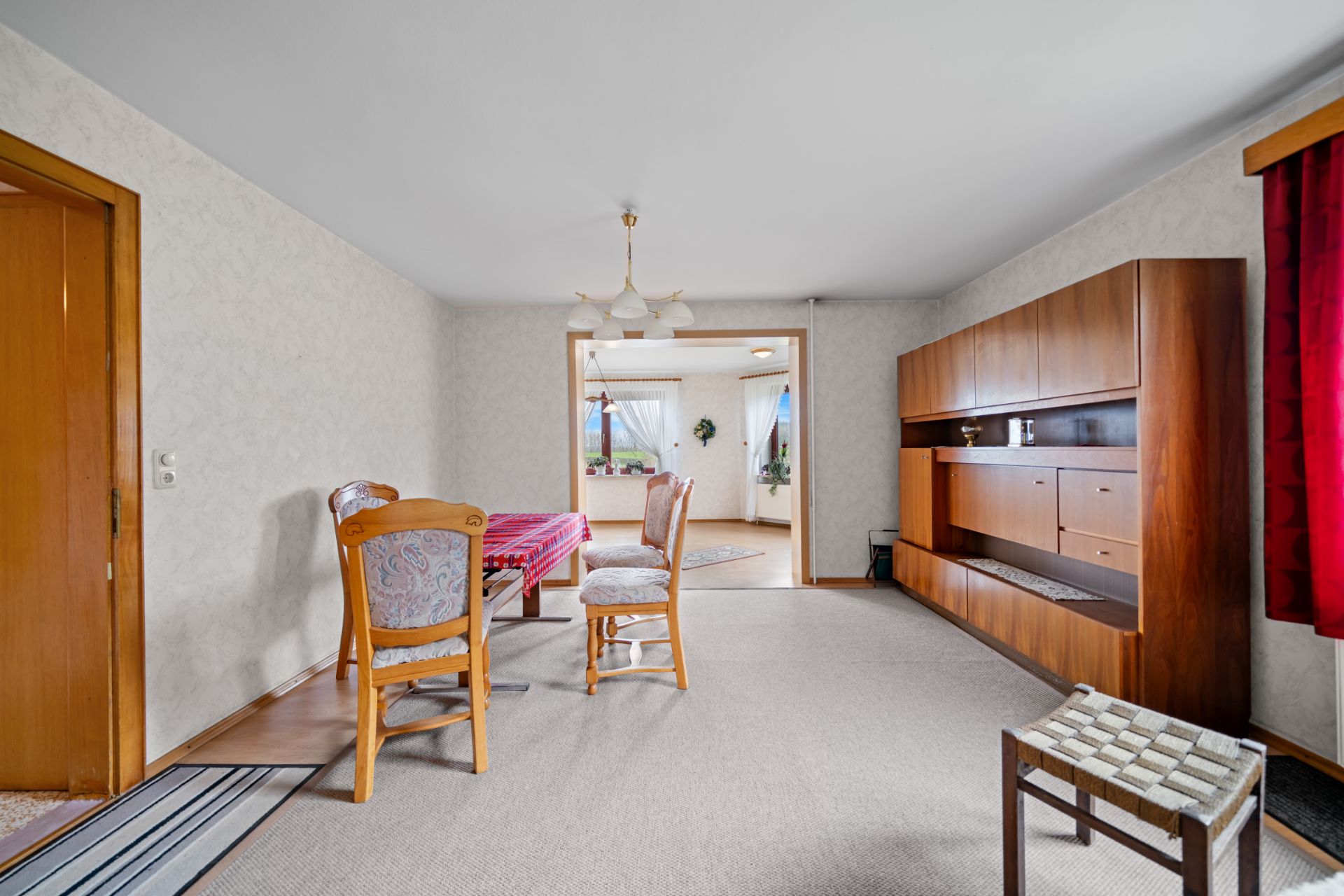 Helles Wohnzimmer mit Esstisch, Stühlen, Holzschrank und rotem Vorhang.