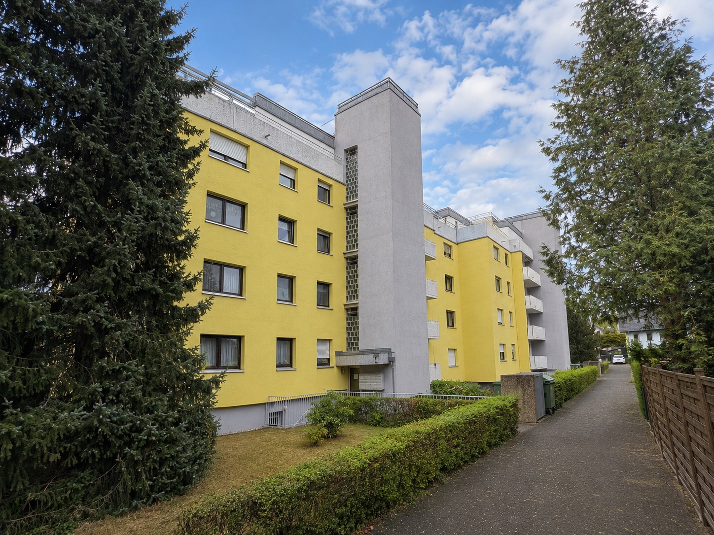 Mehrstöckiges gelbes Wohnhaus mit Balkonen, umgeben von Bäumen und Hecken am Gehweg.