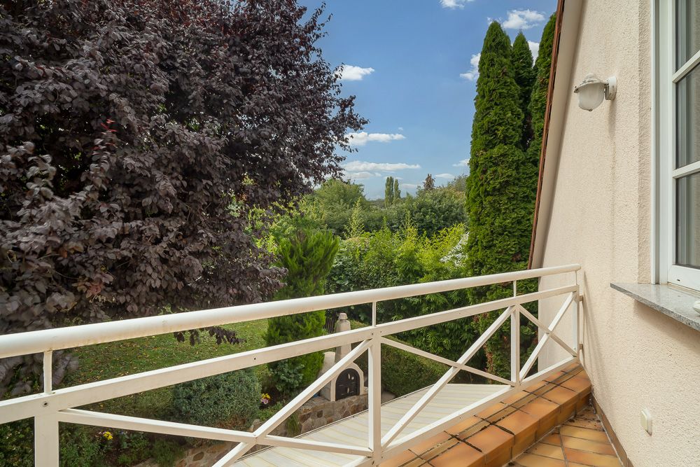 Balkon mit weißem Geländer und Blick auf einen grünen Garten mit Bäumen und Sträuchern.