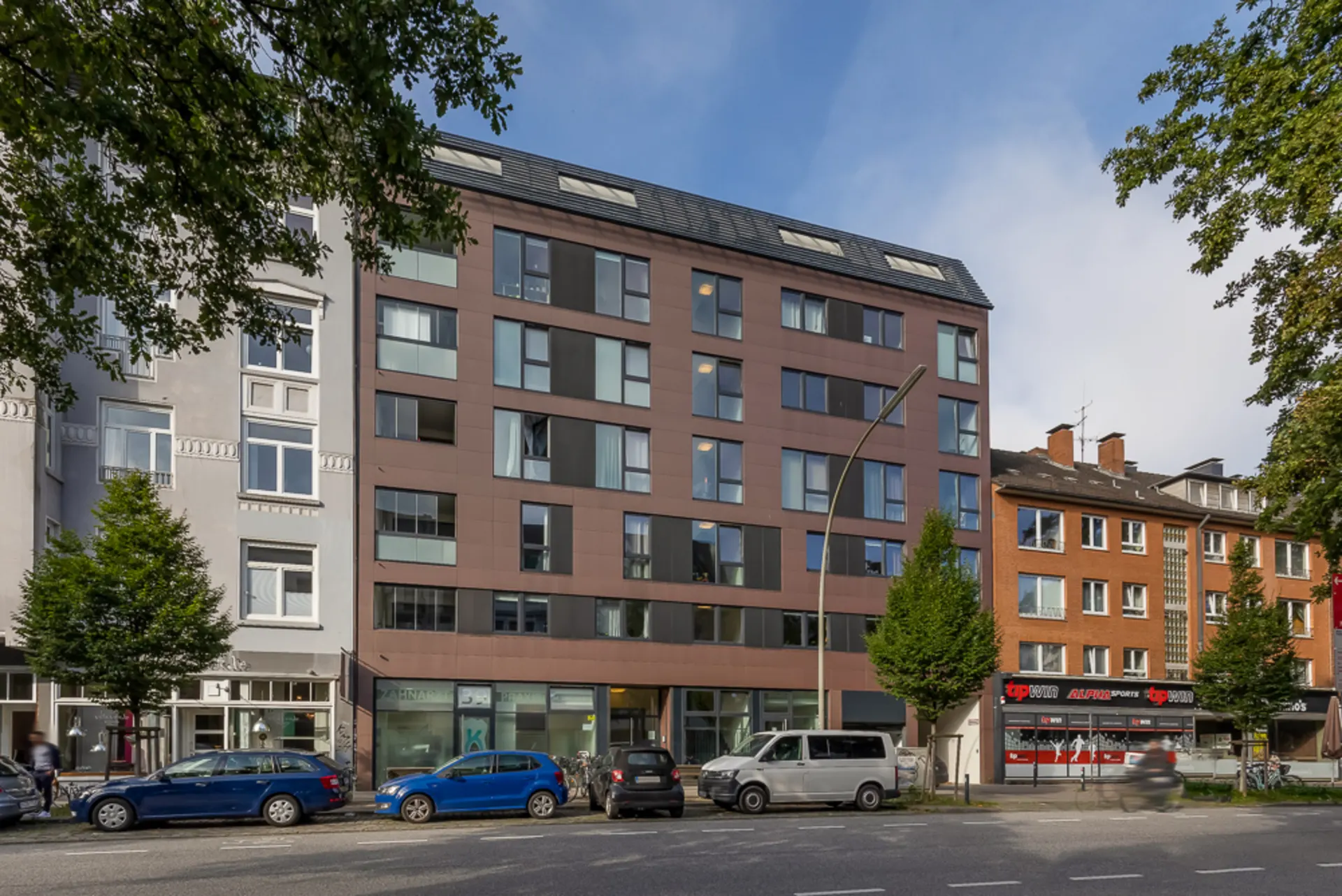 Mehrstöckiges Wohngebäude mit vielen Fenstern an einer Straße, Autos parken davor.