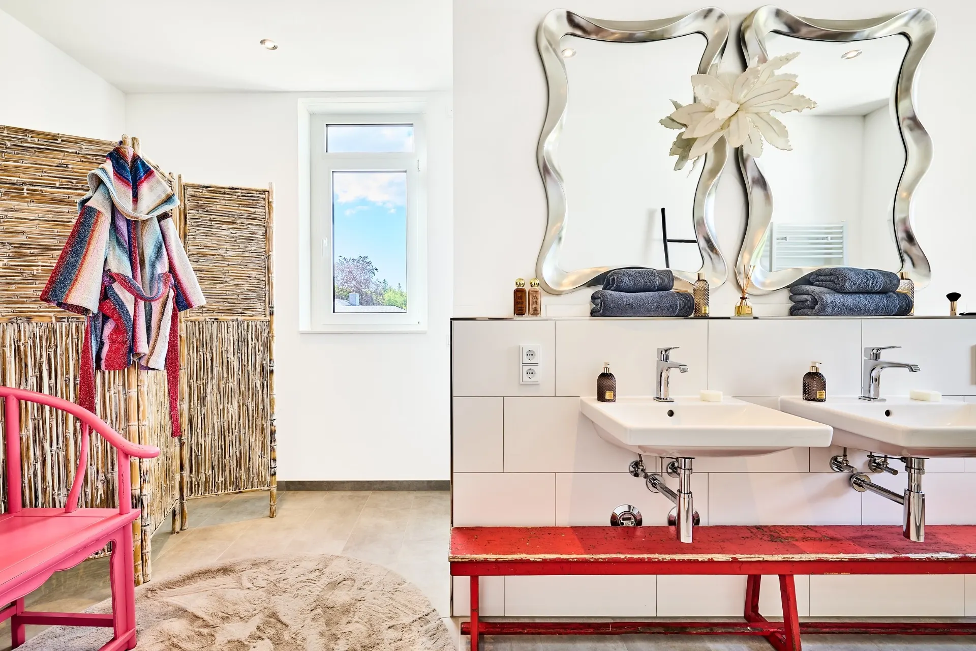 Modernes Badezimmer mit zwei Waschbecken, roten Bank, buntem Bademantel und zwei Spiegeln.