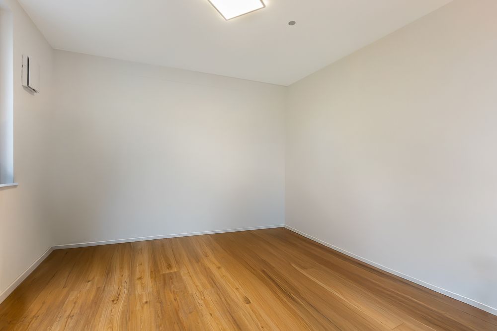 Leerer Raum mit hellen Wänden und Holzboden, Fenster links, Deckenleuchte sichtbar.
